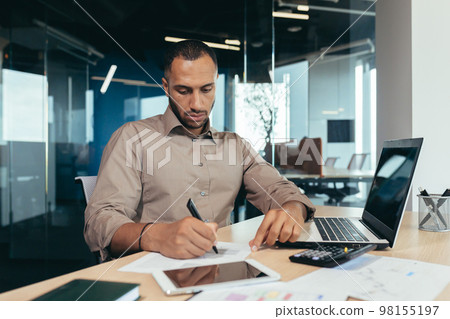 Serious thinking hispanic businessman working with documents and invoices inside office, man behind paper work, using laptop at work sitting at desk. Serious thinking hispanic businessman working with documents and invoices inside office, man behind paper work, using laptop at work sitting at desk. 98155197