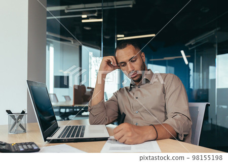 Serious thinking hispanic businessman working with documents and invoices inside office, man behind paper work, using laptop at work sitting at desk. Serious thinking hispanic businessman working with documents and invoices inside office, man behind paper work, using laptop at work sitting at desk. 98155198