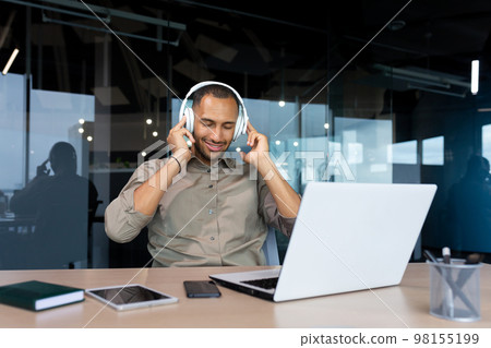 Successful young african american businessman relaxing in office, man in shirt and headphones listening to music online radio and podcasts, sitting at desk using laptop at work. Successful young african american businessman relaxing in office, man in shirt and headphones listening to music online radio and podcasts, sitting at desk using laptop at work. 98155199