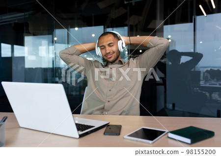 Successful young african american businessman relaxing in office, man in shirt and headphones listening to music online radio and podcasts, sitting at desk using laptop at work. Successful young african american businessman relaxing in office, man in shirt and headphones listening to music online radio and podcasts, sitting at desk using laptop at work. 98155200