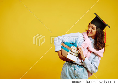 Graduate woman in a graduation hat on her head, with books stands on a yellow background. 98155414