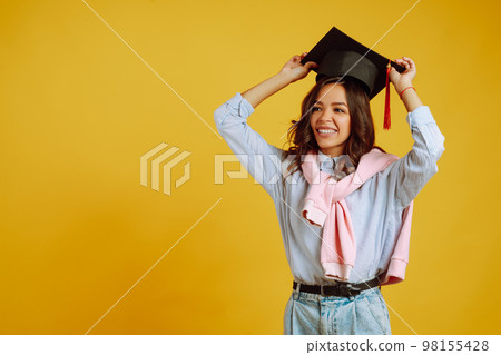 Graduate woman in graduation hat on her head posing on yellow background. Study, education, college. 98155428