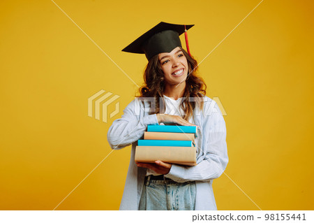 Graduate woman in a graduation hat on her head, with books stands on a yellow background. 98155441