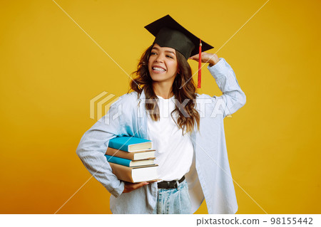 Graduate woman in a graduation hat on her head, with books stands on a yellow background. 98155442