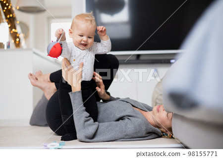Happy family moments. Mother lying comfortably on children's mat playing with her baby boy watching and suppervising his first steps. Positive human emotions, feelings, joy 98155771
