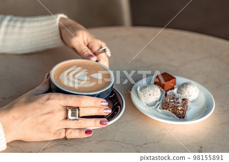 A cup of fresh cappuccino coffee in the hands of a woman on a fashionable background of a white marble table, next to a plate with sweets. Coffee addiction A cup of fresh cappuccino coffee in the hands of a woman on a fashionable background of a white marble table, next to a plate with sweets. Coffee addiction 98155891