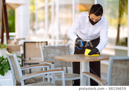 Young Waiter in protective face mask and gloves sanitizing. cleaning the table with disinfectant 98156659