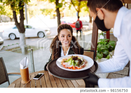 Young waiter in protective face mask and gloves while bringing food to a customer in cafe. Covid-19. Young waiter in protective face mask and gloves while bringing food to a customer in cafe. Covid-19. 98156750