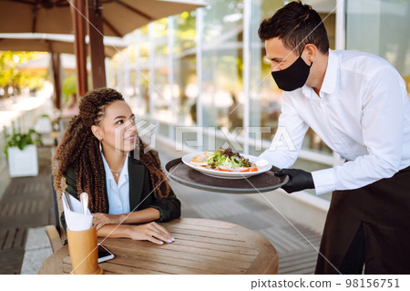 Young waiter in protective face mask and gloves while bringing food to a customer in cafe. Covid-19. Young waiter in protective face mask and gloves while bringing food to a customer in cafe. Covid-19. 98156751