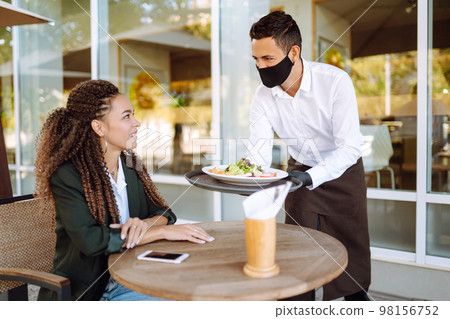 Young waiter in protective face mask and gloves while bringing food to a customer in cafe. Covid-19. Young waiter in protective face mask and gloves while bringing food to a customer in cafe. Covid-19. 98156752