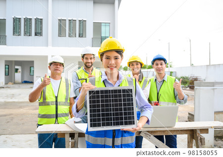 electrician woman in safety vest with helmet showing solar cell panel with the team in background, standing in the under-construction building. Green energy electricity concept. concept of sustainable 98158055