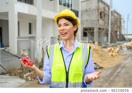 Young construction engineer woman in safety vest with yellow helmet holding radio, standing on building construction site. Smiling talking to the camera. 98158056