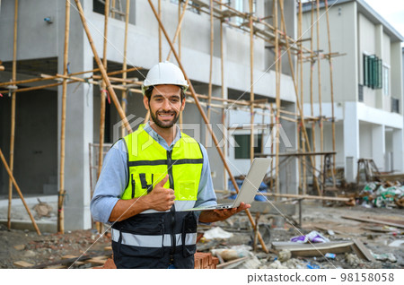 Young attractive construction smiling bearded in vest with white helmet holding laptop and showing thumbs up gesture while standing on construction site. Home building project. Young attractive construction smiling bearded in vest with white helmet holding laptop and showing thumbs up gesture while standing on construction site. Home building project. 98158058