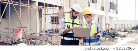 Young attractive construction man and woman in vest with helmet working with laptop, standing on building construction site. Home building project. Engineer foreman discusses with a coworker at Young attractive construction man and woman in vest with helmet working with laptop, standing on building construction site. Home building project. Engineer foreman discusses with a coworker at 98158060