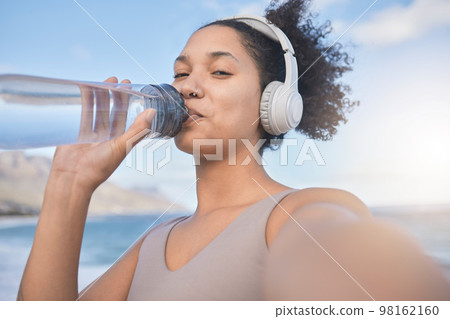 Woman runner, drinking water and selfie with music while outdoor for exercise, workout or fitness. Black woman, headphones and beach for training, wellness or health in nature by ocean in summer sun 98162160
