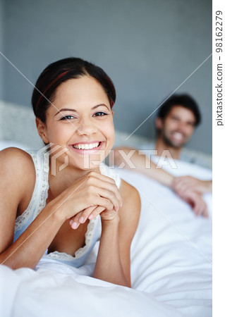 I love lying in. Portrait of a smiling woman lying on her bed with her boyfriend in the background. 98162279