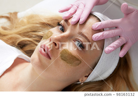 Cosmetologist applying a clay mask to the face of beautiful woman. Spa treatment and face care in the beauty salon Cosmetologist applying a clay mask to the face of beautiful woman. Spa treatment and face care in the beauty salon 98163132