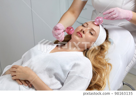 Young woman receiving facial massage with glass balls in beauty salon Young woman receiving facial massage with glass balls in beauty salon 98163135