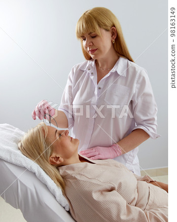 Woman getting her eyebrows angled by a beautician in a beauty salon with instrument Woman getting her eyebrows angled by a beautician in a beauty salon with instrument 98163149