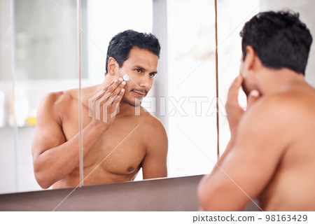 Being this sexy takes hard work. A young man applying cream to his face while looking in the mirror. 98163429