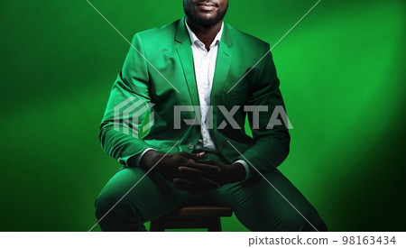Fashion, suit and black man model with a formal outfit sitting on a chair while posing in a studio. Elegant, trendy and African male in luxury, stylish and fancy clothes isolated by green background. Fashion, suit and black man model with a formal outfit sitting on a chair while posing in a studio. Elegant, trendy and African male in luxury, stylish and fancy clothes isolated by green background. 98163434