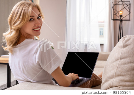 Woman on sofa working on laptop with mockup blank screen. Empty copy space on monitor for advertisement. Black screen. 98165504