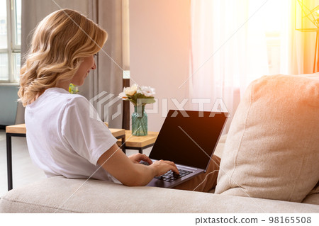 Woman on sofa working on laptop with mockup blank screen. Empty copy space on monitor for advertisement. Black screen. 98165508