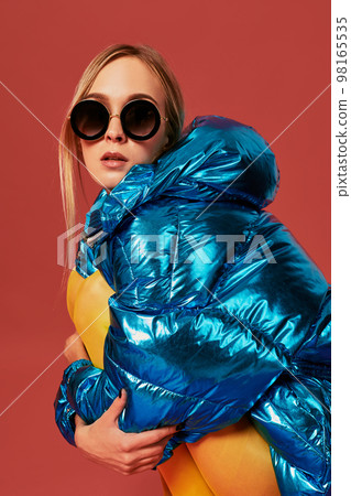 Young cute nice blonde girl in blue jacket sits in a fetal position in studio before camera, red background. Side view Young cute nice blonde girl in blue jacket sits in a fetal position in studio before camera, red background. Side view 98165535