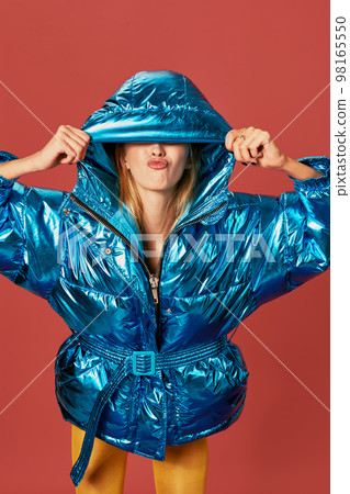Blonde slim girl in blue down jacket covers her face from wind with hood Blonde slim girl in blue down jacket covers her face from wind with hood 98165550