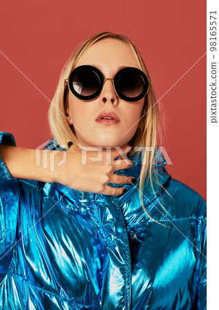 Concept art image of slim blonde girl in studio, wearing blue down jacket, posing on camera. 98165571