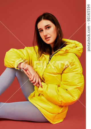 Studio portrait of young brunette woman sitting on the floor in yellow down jacket and grey blue panty hoses or stockings 98165594