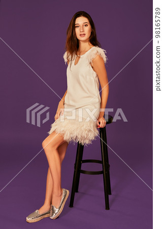 fashion photo of beautiful woman with brunette hair in white dress posing on chair at studio 98165789