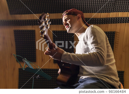 Caucasian man sings song, playing guitar, holding musical instrument in hands, sitting in chair in studio. Darken photo. Male wears casual cloth. Hobby, leisure of creative person, artist. Horizontal 98166484