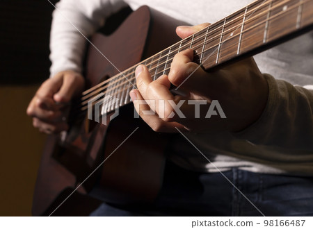 Closeup white man plays guitar, holding musical instrument in hands, sitting on chair in studio. Darken photo. Male wears casual cloth. Hobby, leisure of creative person, artist. Horizontal 98166487