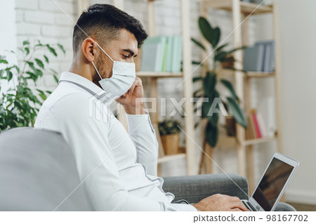 Young businessman in face mask sitting on sofa and using laptop 98167702