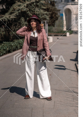 Woman park city. Stylish woman in a hat walks in a park in the city. Dressed in white corset trousers and a pink jacket with a bag in her hands. Woman park city. Stylish woman in a hat walks in a park in the city. Dressed in white corset trousers and a pink jacket with a bag in her hands. 98167909