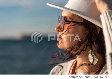 Portrait of a curly haired woman in a white hat and glasses on the background of the sea. Vacation on the sea, walk, tourism 98167915