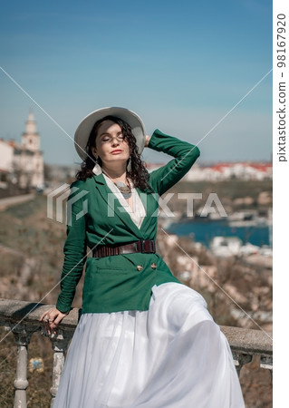 Woman walks around the city, lifestyle. A young beautiful woman in a green jacket, white skirt and hat is sitting on a white fence with balusters overlooking the sea bay and the city. Woman walks around the city, lifestyle. A young beautiful woman in a green jacket, white skirt and hat is sitting on a white fence with balusters overlooking the sea bay and the city. 98167920