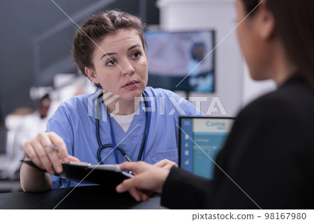 Nurse showing patient expertise to receptionist discussing disease symptoms while asking her to programming another examination. Medical staff working in hospital waiting area, health care support 98167980