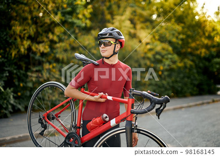 Sporty male in cycling clothes, protective helmet and glasses carrying bike, posing 98168145