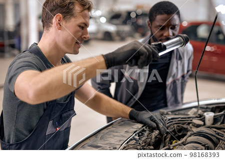 two diverse mechanics checking car hood in auto service, cooperate, work together two diverse mechanics checking car hood in auto service, cooperate, work together 98168393