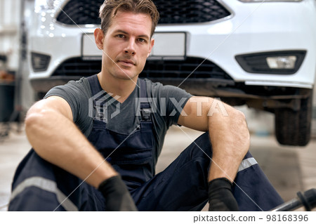 At auto service. Handsome young auto mechanic in uniform sit on floor having rest 98168396