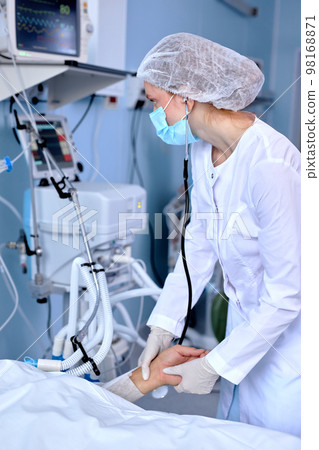 Experienced Female Doctor Checking The Pulse Of Patient Lying On Bed, Holding Hands Experienced Female Doctor Checking The Pulse Of Patient Lying On Bed, Holding Hands 98168871
