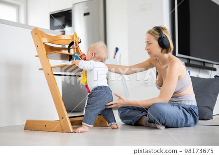 Women's multitasking. Mother sitting on floor playing with her baby boy watching and suppervising his first steps while listening to podcast on wireless headphones Women's multitasking. Mother sitting on floor playing with her baby boy watching and suppervising his first steps while listening to podcast on wireless headphones 98173675