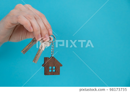 A woman's hand holds the keys with a keychain in the shape of a house on a blue background.  98173735