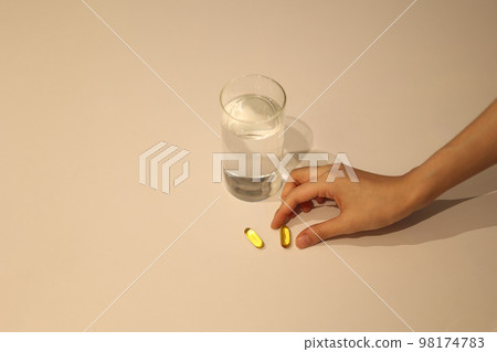 Woman's hand holding a pill and a glass of water on a white table Woman's hand holding a pill and a glass of water on a white table 98174783