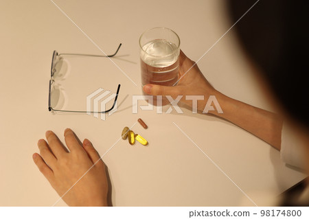 Pills, glass of water and glasses are placed on a white table, a woman holding a glass of water looking at the medicine 98174800