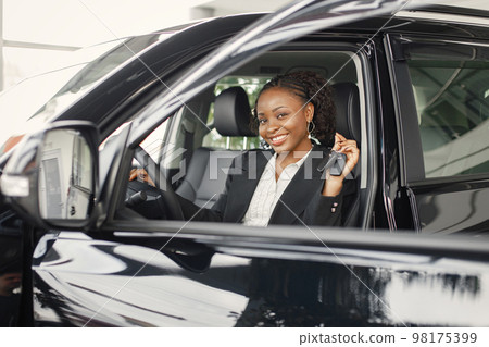 Young and cheerful woman enjoying new car while sitting inside. Black woman holding a car key. Girl wearing black costume. 98175399