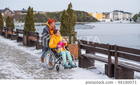A woman in a wheelchair walks with her friend and a dog by the lake in winter. 98180071