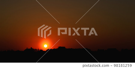 The setting sun at sunset in the orange evening sky above the silhouettes of buildings and trees 98181259